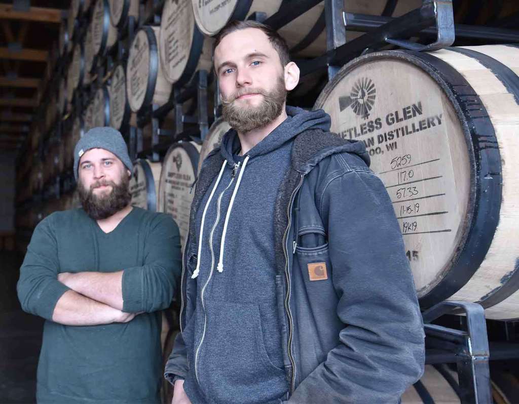 Distillers Nathan and Max pose in the rackhouse in front of barrels of aging spirits. 