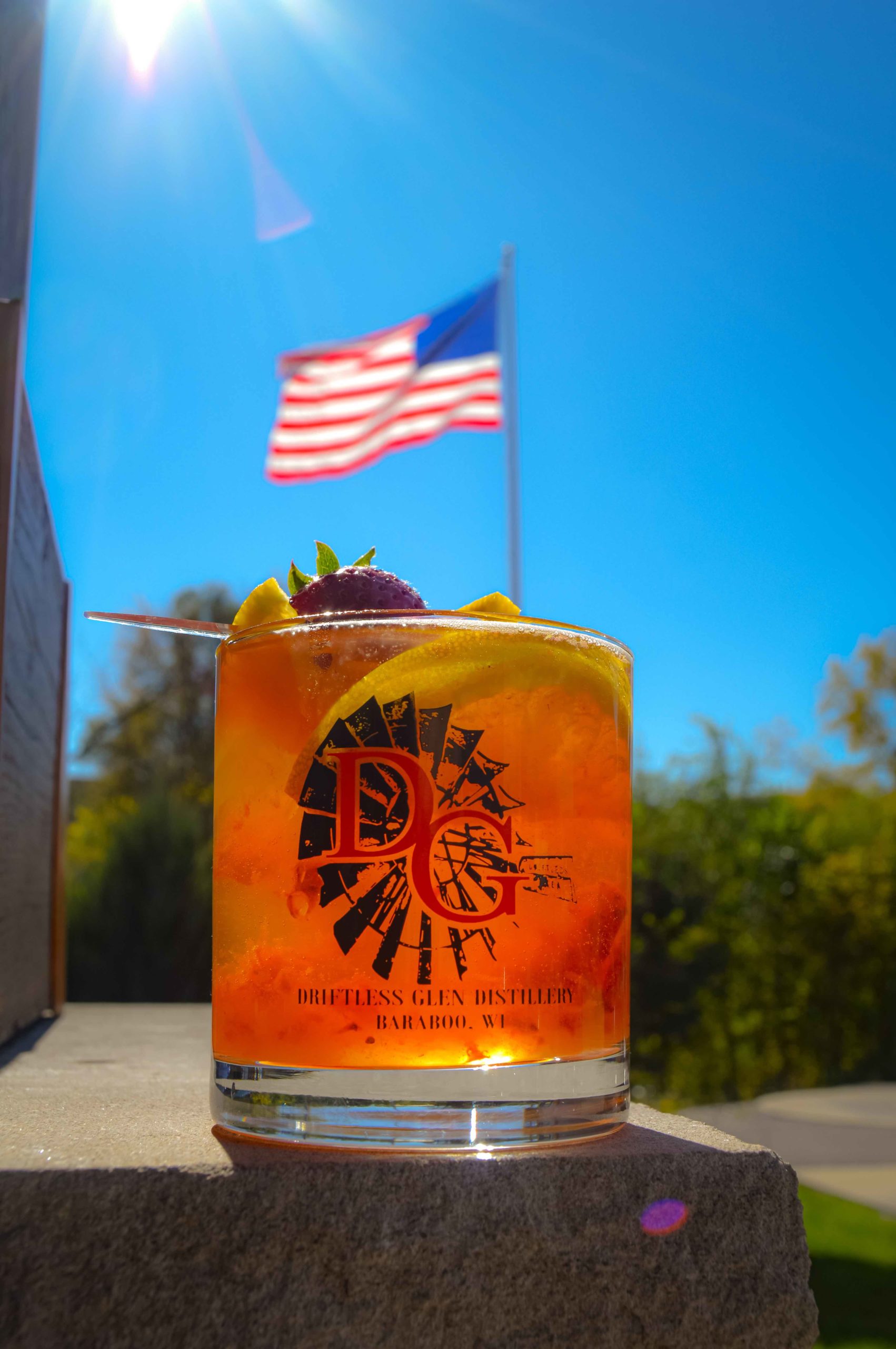 Strawberry Peach Old Fashioned for #mixologymonday sits in front of a blue sky outside with the flag pole in the background